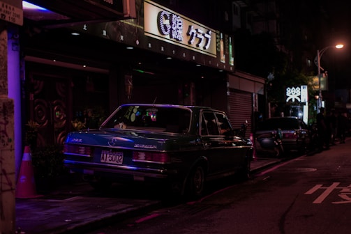 A moody urban night scene with Johnny Kaikkllo walking past glowing city lights and a classic Mercedes parked nearby.