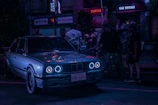 A group of car enthusiasts gathered around a neon-lit street corner, sharing music and stories.