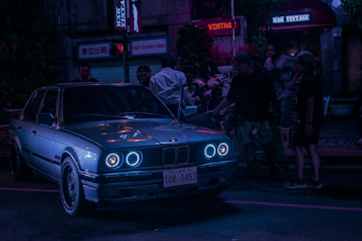 A group of car enthusiasts gathered around a neon-lit street corner, sharing music and stories.