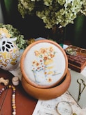 A cozy and artistic workspace featuring a cross-stitch embroidery of flowers in pots, displayed on a wooden hoop. The setting is adorned with a decorative ceramic item, sheet music, vintage spectacles, and a pair of small scissors. Green hydrangea flowers add a natural touch in the background, while wooden stamps and colorful beads suggest a creative environment.