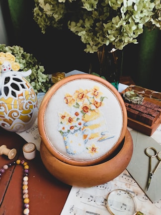 A cozy and artistic workspace featuring a cross-stitch embroidery of flowers in pots, displayed on a wooden hoop. The setting is adorned with a decorative ceramic item, sheet music, vintage spectacles, and a pair of small scissors. Green hydrangea flowers add a natural touch in the background, while wooden stamps and colorful beads suggest a creative environment.