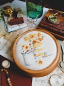 An assortment of objects placed on a table, including a hand-embroidered fabric showing a design of two pitchers with yellow and orange flowers, a green glass bottle, vintage papers and envelopes, a wooden stamp, a pair of glasses, a beaded bracelet, and a small gold-colored box. There are also other small items like scissors, a candle, and a wooden tray with a numerical stamp set.