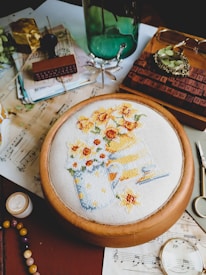 An assortment of objects placed on a table, including a hand-embroidered fabric showing a design of two pitchers with yellow and orange flowers, a green glass bottle, vintage papers and envelopes, a wooden stamp, a pair of glasses, a beaded bracelet, and a small gold-colored box. There are also other small items like scissors, a candle, and a wooden tray with a numerical stamp set.