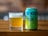A glass filled with a pale, golden beer sits on a wooden surface next to a colorful beer can. The can features bold lettering and vibrant colors, with branding details prominently displayed.