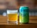 A glass filled with a pale, golden beer sits on a wooden surface next to a colorful beer can. The can features bold lettering and vibrant colors, with branding details prominently displayed.