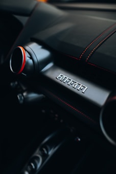Close-up view of a car dashboard featuring red stitching on black leather and a logo emblem on a sleek metallic panel. The design of the knob and buttons suggests a premium vehicle interior.