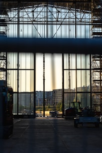 A large industrial hall with tall glass walls and scaffolding on either side, looking out onto an urban landscape with buildings in the distance. Construction vehicles are visible in the foreground, and the lighting suggests either early morning or late afternoon.