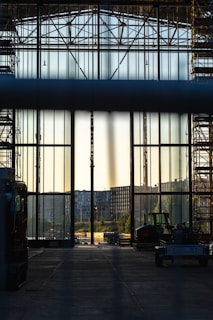 A large industrial hall with tall glass walls and scaffolding on either side, looking out onto an urban landscape with buildings in the distance. Construction vehicles are visible in the foreground, and the lighting suggests either early morning or late afternoon.