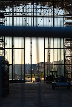 A large industrial hall with tall glass walls and scaffolding on either side, looking out onto an urban landscape with buildings in the distance. Construction vehicles are visible in the foreground, and the lighting suggests either early morning or late afternoon.