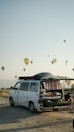 A camper van with its rear door open reveals a cozy interior setup, including a bed. The van is parked on a sandy or gravel surface, with a backdrop of hot air balloons scattered across the sky. The scene conveys a sense of adventure and travel, set against a clear sky during early morning or late afternoon. A person can be seen sitting outside, absorbed in the relaxing atmosphere.