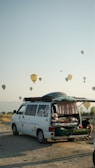 A camper van with its rear door open reveals a cozy interior setup, including a bed. The van is parked on a sandy or gravel surface, with a backdrop of hot air balloons scattered across the sky. The scene conveys a sense of adventure and travel, set against a clear sky during early morning or late afternoon. A person can be seen sitting outside, absorbed in the relaxing atmosphere.