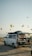 A camper van with its rear door open reveals a cozy interior setup, including a bed. The van is parked on a sandy or gravel surface, with a backdrop of hot air balloons scattered across the sky. The scene conveys a sense of adventure and travel, set against a clear sky during early morning or late afternoon. A person can be seen sitting outside, absorbed in the relaxing atmosphere.