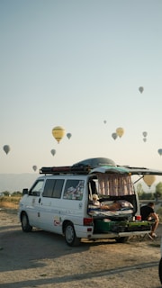 A camper van with its rear door open reveals a cozy interior setup, including a bed. The van is parked on a sandy or gravel surface, with a backdrop of hot air balloons scattered across the sky. The scene conveys a sense of adventure and travel, set against a clear sky during early morning or late afternoon. A person can be seen sitting outside, absorbed in the relaxing atmosphere.