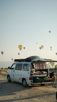A camper van with its rear door open reveals a cozy interior setup, including a bed. The van is parked on a sandy or gravel surface, with a backdrop of hot air balloons scattered across the sky. The scene conveys a sense of adventure and travel, set against a clear sky during early morning or late afternoon. A person can be seen sitting outside, absorbed in the relaxing atmosphere.