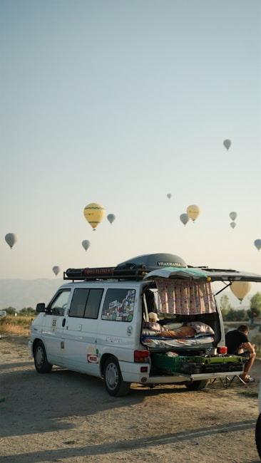 A camper van with its rear door open reveals a cozy interior setup, including a bed. The van is parked on a sandy or gravel surface, with a backdrop of hot air balloons scattered across the sky. The scene conveys a sense of adventure and travel, set against a clear sky during early morning or late afternoon. A person can be seen sitting outside, absorbed in the relaxing atmosphere.