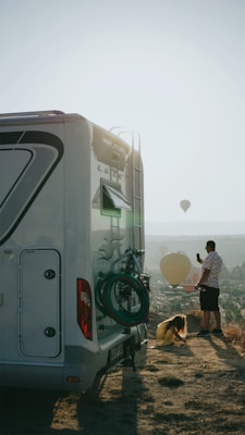 A person stands beside a large recreational vehicle (RV), taking a photo with their phone, while another person crouches nearby. In the background, several hot air balloons are in the sky over a landscape that includes hills and some structures.