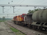 A freight train is stationed on the railway track, consisting of multiple cargo cars. The train has a red and yellow locomotive at the front. Overhead electric lines span across the rails, and there is greenery and trees in the background. A concrete platform is visible to the left, with a rail track running parallel to it.