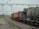A freight train is stationed on the railway track, consisting of multiple cargo cars. The train has a red and yellow locomotive at the front. Overhead electric lines span across the rails, and there is greenery and trees in the background. A concrete platform is visible to the left, with a rail track running parallel to it.