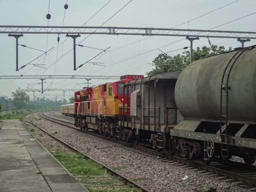 A freight train is stationed on the railway track, consisting of multiple cargo cars. The train has a red and yellow locomotive at the front. Overhead electric lines span across the rails, and there is greenery and trees in the background. A concrete platform is visible to the left, with a rail track running parallel to it.