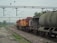A freight train is stationed on the railway track, consisting of multiple cargo cars. The train has a red and yellow locomotive at the front. Overhead electric lines span across the rails, and there is greenery and trees in the background. A concrete platform is visible to the left, with a rail track running parallel to it.