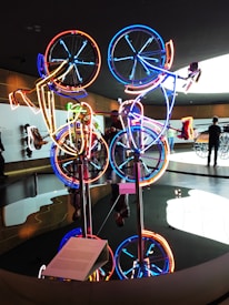 An artistic installation featuring a series of bicycles with neon lights outlining their frames and wheels in various bright colors. The display is situated in a modern gallery space with people observing the artwork. The reflection of the installation is visible on the glossy surface below, creating a vivid, mirrored effect.