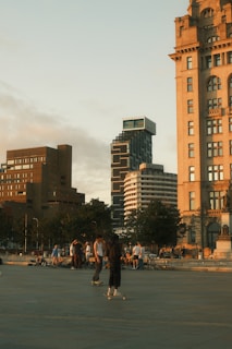 A group of people are skateboarding in an urban area with modern high-rise buildings and historical architecture in the background. The scene is captured during golden hour, creating a warm and relaxed atmosphere. Trees and scattered benches are present around the square, and a statue can be seen on the right side.