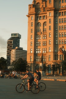 A group of people are seen enjoying a leisurely bike ride near an historic building in a city setting. The architecture is grand and old, contrasted with modern buildings in the background. The warm glow of the setting sun casts a serene light over the entire scene.