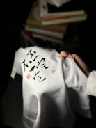 A hand holding a white garment with black calligraphic writing and red stamps. The writing and stamps are prominently displayed on the fabric. In the background, blurred piles of books add context to the setting.