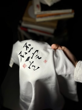 A hand holding a white garment with black calligraphic writing and red stamps. The writing and stamps are prominently displayed on the fabric. In the background, blurred piles of books add context to the setting.