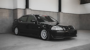 A sleek black sedan with tinted windows is parked indoors on a light gray tiled floor. The car features white rims and has a sporty appearance, accentuated by its smooth lines and low stance. The background consists of gray walls with subtle panel detailing, creating a minimalist and clean setting.