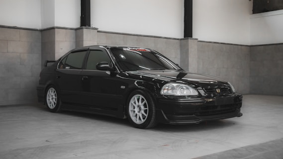 A sleek black sedan with tinted windows is parked indoors on a light gray tiled floor. The car features white rims and has a sporty appearance, accentuated by its smooth lines and low stance. The background consists of gray walls with subtle panel detailing, creating a minimalist and clean setting.