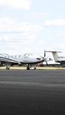 A small private jet is parked on an airfield, positioned on a paved surface with grassy areas in the background. The sky is mostly cloudy with some patches of blue.