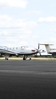 A small private jet is parked on an airfield, positioned on a paved surface with grassy areas in the background. The sky is mostly cloudy with some patches of blue.