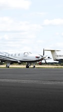 A small private jet is parked on an airfield, positioned on a paved surface with grassy areas in the background. The sky is mostly cloudy with some patches of blue.