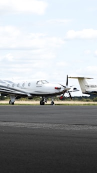 A small private jet is parked on an airfield, positioned on a paved surface with grassy areas in the background. The sky is mostly cloudy with some patches of blue.
