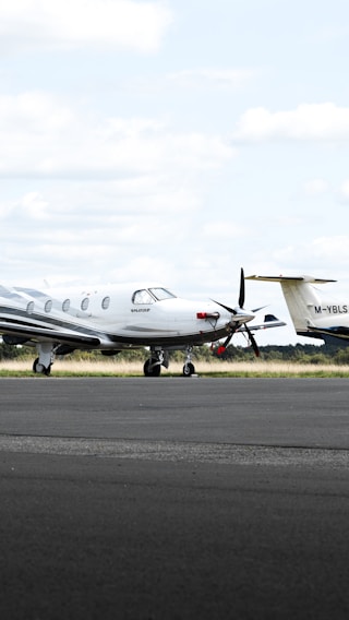 A small private jet is parked on an airfield, positioned on a paved surface with grassy areas in the background. The sky is mostly cloudy with some patches of blue.