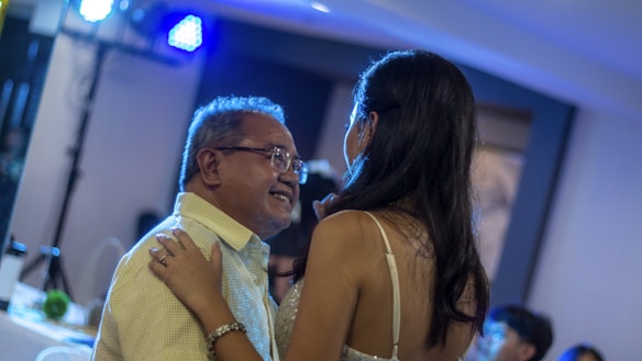 An older man and a younger woman are sharing a dance at what seems to be a social gathering or celebration. The man is smiling warmly, and the woman has her arms around his shoulders. The setting appears intimate with dim lighting and a soft blue glow from overhead lights.