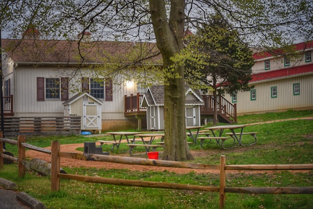 A quaint rural scene depicts several wooden buildings with red roofs, surrounded by greenery. In the foreground, a large tree stands near picnic tables and a wooden fence. The ground is lined with gravel paths leading to the small buildings.