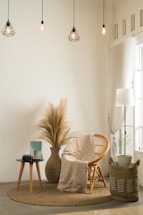 A cozy and minimalist living space featuring a wicker chair with a patterned cream throw draped over it. A round indoor rug lies beneath the chair. Next to it, a side table holds a camera and a book. A large vase with dried pampas grass adds a natural element to the decor. Hanging above are geometric pendant lamps with warm lighting, and a tall lamp sits beside sheer curtains by the window. A woven basket with a potted plant adds texture to the corner.