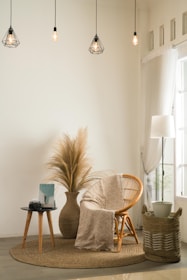 A cozy and minimalist living space featuring a wicker chair with a patterned cream throw draped over it. A round indoor rug lies beneath the chair. Next to it, a side table holds a camera and a book. A large vase with dried pampas grass adds a natural element to the decor. Hanging above are geometric pendant lamps with warm lighting, and a tall lamp sits beside sheer curtains by the window. A woven basket with a potted plant adds texture to the corner.