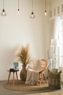 A cozy and minimalist living space featuring a wicker chair with a patterned cream throw draped over it. A round indoor rug lies beneath the chair. Next to it, a side table holds a camera and a book. A large vase with dried pampas grass adds a natural element to the decor. Hanging above are geometric pendant lamps with warm lighting, and a tall lamp sits beside sheer curtains by the window. A woven basket with a potted plant adds texture to the corner.
