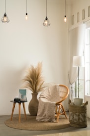 A cozy and minimalist living space featuring a wicker chair with a patterned cream throw draped over it. A round indoor rug lies beneath the chair. Next to it, a side table holds a camera and a book. A large vase with dried pampas grass adds a natural element to the decor. Hanging above are geometric pendant lamps with warm lighting, and a tall lamp sits beside sheer curtains by the window. A woven basket with a potted plant adds texture to the corner.
