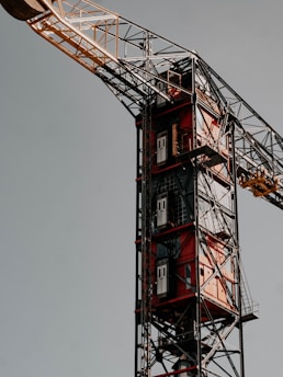 A close-up view of a construction crane with a red and white color scheme. The crane's metallic structure is complex, featuring a vertical lift system with multiple levels and doors. The sky in the background is a muted gray, casting the structure in a light that highlights its intricate details and robust framework.