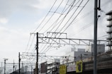 Complex network of overhead railway wires stretches across the sky with a backdrop of urban buildings. Several utility poles support the wires, and signs with height restrictions are visible below. The sky above is partly cloudy, adding a sense of openness.