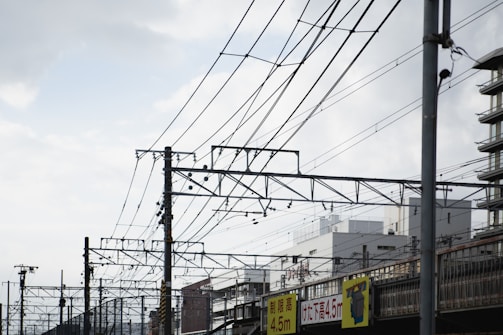 Complex network of overhead railway wires stretches across the sky with a backdrop of urban buildings. Several utility poles support the wires, and signs with height restrictions are visible below. The sky above is partly cloudy, adding a sense of openness.