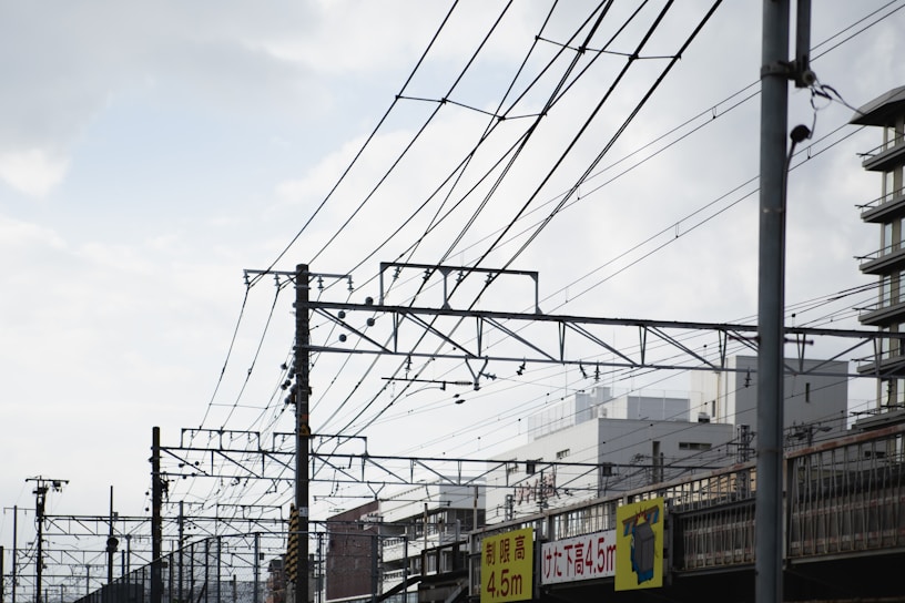 Complex network of overhead railway wires stretches across the sky with a backdrop of urban buildings. Several utility poles support the wires, and signs with height restrictions are visible below. The sky above is partly cloudy, adding a sense of openness.