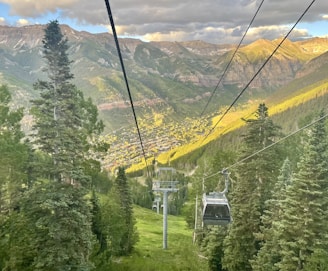 A mountainous landscape featuring a gondola lift traveling over a lush, green forest. In the distance, a town is nestled within the valley surrounded by towering mountains. The sky is partly cloudy, with sunlight casting warm highlights on the peaks.
