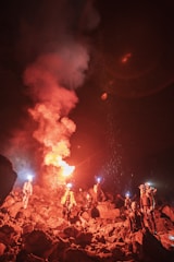 A group of people is gathered on rocky terrain at night, holding lights and flares that illuminate the surroundings with a dramatic glow. The rocky landscape is rugged, and the people appear to be wearing outdoor gear, possibly for hiking or spelunking.