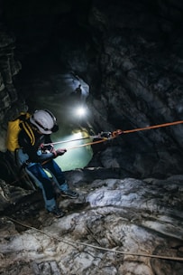 A person is engaged in a caving or rappelling activity inside a rocky cave. They are equipped with climbing gear including a helmet and ropes. The cave is dark, illuminated by the light from the person, highlighting the rugged textures of the stones and the presence of a body of water in the background.