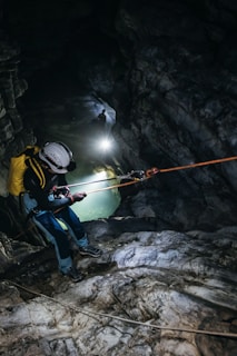 A person is engaged in a caving or rappelling activity inside a rocky cave. They are equipped with climbing gear including a helmet and ropes. The cave is dark, illuminated by the light from the person, highlighting the rugged textures of the stones and the presence of a body of water in the background.